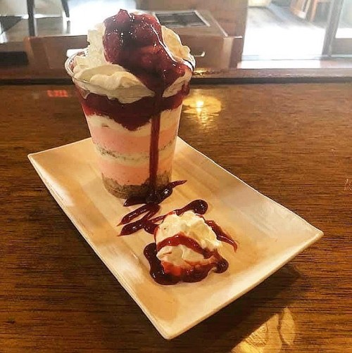 A layered dessert in a glass with pink and white cream, topped with whipped cream and berry sauce, served on a plate.