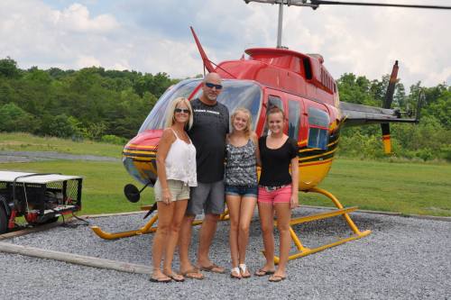 A group of four people poses in front of a red and yellow helicopter on a grassy field.