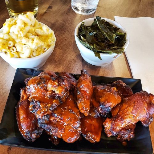 A plate of glazed chicken wings with sides of macaroni and cheese and collard greens on a wooden table.