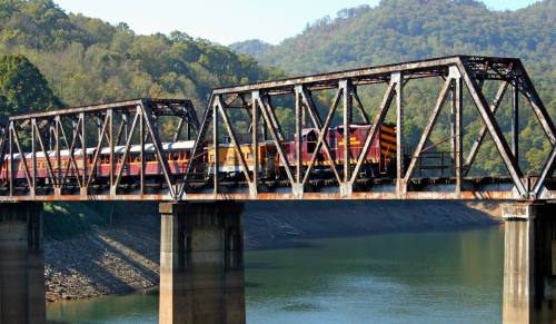 A colorful train crosses a rusty metal bridge over a calm river, surrounded by green hills.