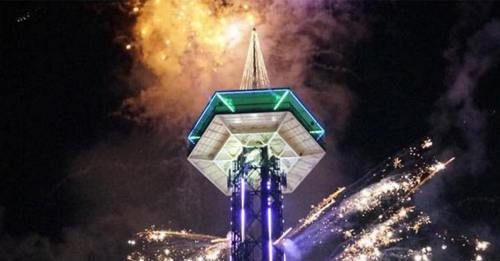 Fireworks illuminate the night sky around a tall tower with a green and blue lit structure.