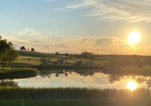 A serene landscape featuring a pond reflecting the sunset, surrounded by lush greenery and distant hills.
