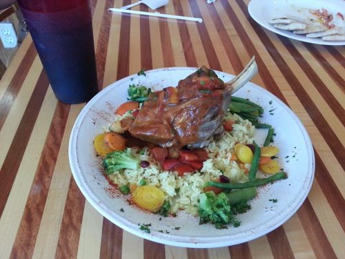 A plate of rice topped with a large lamb chop, mixed vegetables, and a drink on a wooden table.