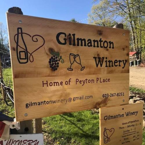 Sign for Gilmanton Winery featuring a wine bottle, grapes, and glasses, with contact info and hours of operation.