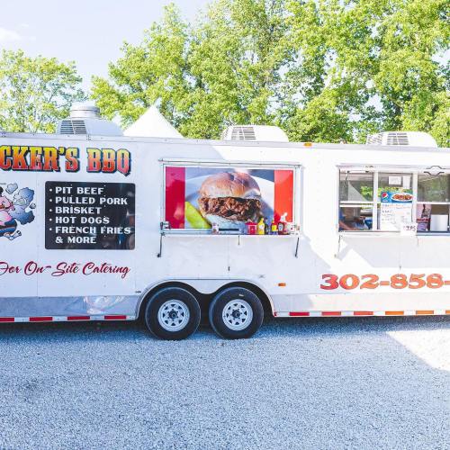 A food truck named "Pucker's BBQ" serving BBQ dishes like brisket, pulled pork, and hot dogs in a sunny outdoor setting.