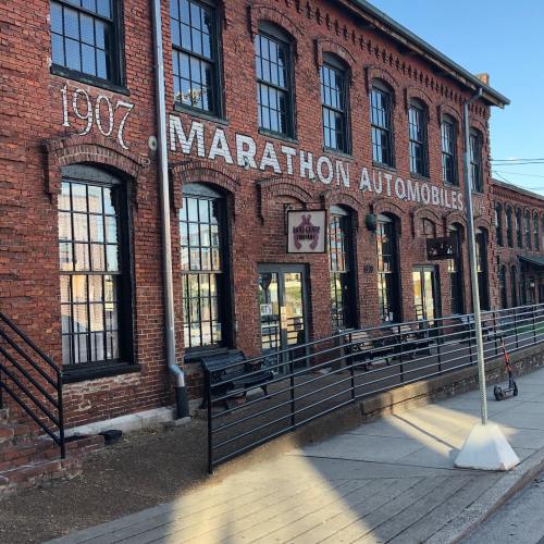 Historic brick building with large windows, featuring "Marathon Automobiles" signage, set in a sunny outdoor area.