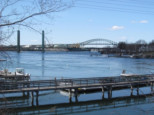 A view of a river with two bridges in the background and a wooden dock in the foreground on a clear day.