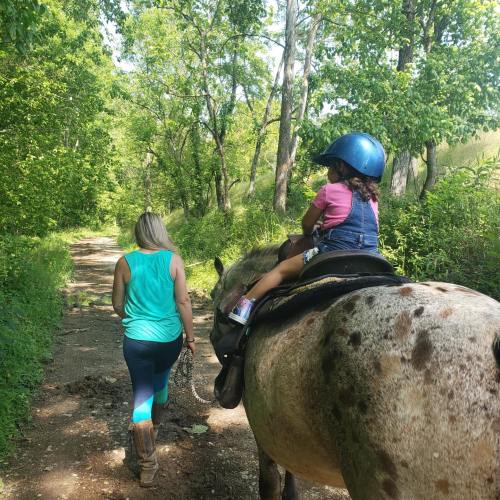 A woman leads a horse along a wooded path, with a child riding on the horse, both enjoying a sunny day outdoors.