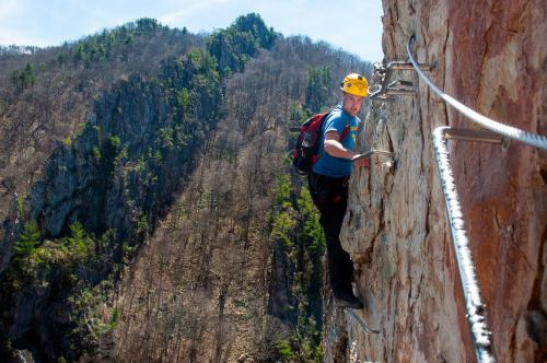A climber ascends a rocky cliff using safety cables, surrounded by a mountainous landscape.