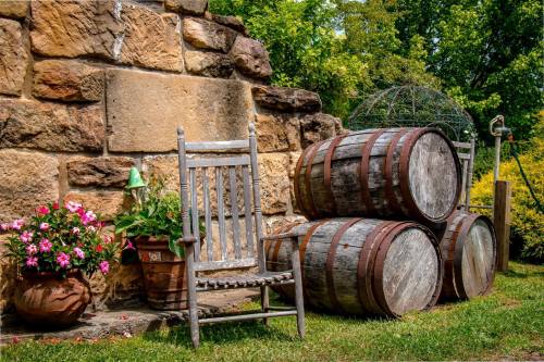 A rustic scene featuring a wooden rocking chair, stacked barrels, and potted flowers against a stone wall.