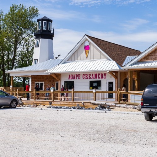 A charming ice cream shop named Agape Creamery with a lighthouse design, surrounded by a gravel parking lot and visitors.