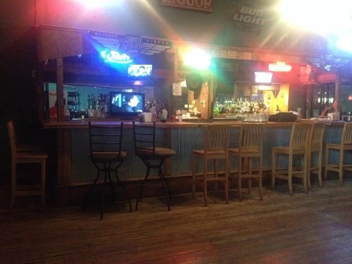 A dimly lit bar with a wooden counter, empty stools, and colorful neon lights reflecting off the walls.