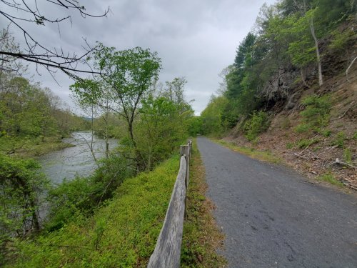 A scenic path alongside a river, surrounded by lush greenery and trees under a cloudy sky.