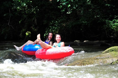 Two friends relax on colorful inner tubes in a river, smiling and enjoying a sunny day surrounded by greenery.