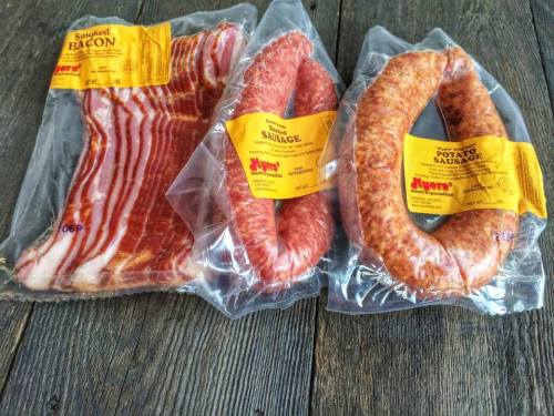Three packages of meat: smoked bacon, sausage, and potato sausage, all sealed in plastic on a wooden surface.