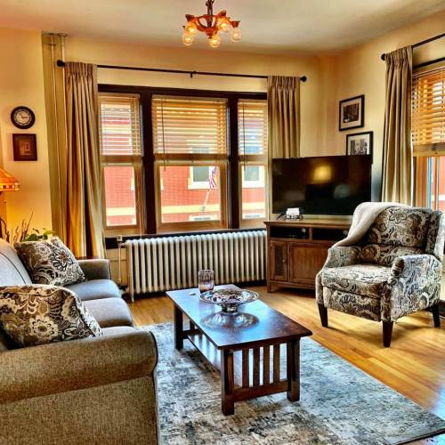 Cozy living room with a sofa, patterned armchair, coffee table, and a TV, featuring large windows and warm lighting.