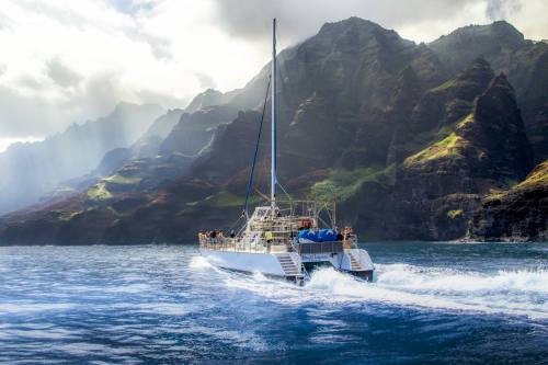 A catamaran sails through blue waters near dramatic, mountainous coastline under a cloudy sky.