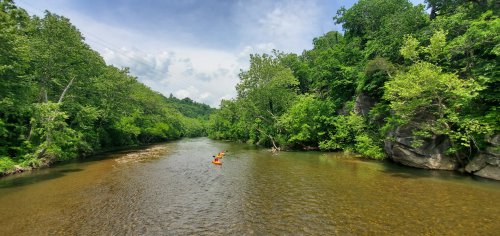 A kayaker paddles down a calm river surrounded by lush green trees and rocky banks under a partly cloudy sky.
