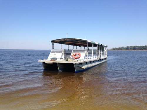 A flat-bottomed boat anchored on calm water, with a clear blue sky and trees in the background.