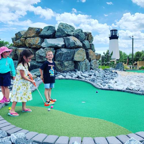 Three children play mini-golf on a green course with rocks and a lighthouse in the background under a blue sky.
