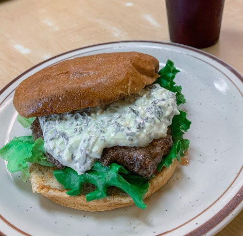A hamburger with lettuce and creamy sauce on a whole grain bun, served on a plate with a drink in the background.