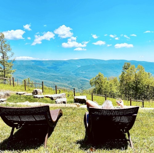 A person relaxes in a lounge chair, overlooking a scenic mountain view under a clear blue sky.