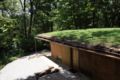 A unique earth-sheltered home with a grassy roof, surrounded by lush trees and a gravel pathway.