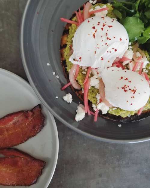 A plate featuring avocado toast topped with poached eggs, pickled onions, feta, and a side of crispy bacon.