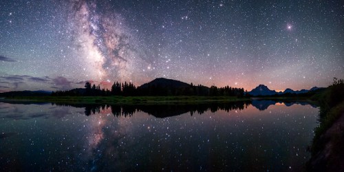 A stunning night sky filled with stars and the Milky Way, reflecting on a calm lake surrounded by mountains and trees.