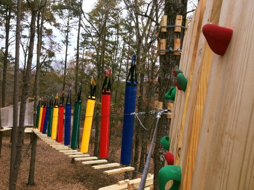 A colorful rope course with climbing holds and hanging cylindrical obstacles among trees in a forested area.