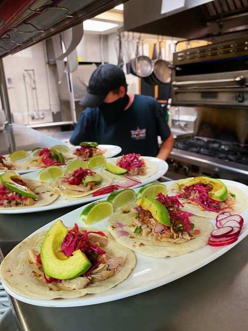 A chef prepares plates of tacos topped with avocado, lime, and pickled vegetables in a busy kitchen.