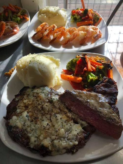 A plate featuring a steak with melted cheese, mashed potatoes, shrimp, and a colorful vegetable medley.