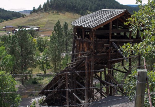 A weathered wooden structure on a hillside, surrounded by trees and fields, with a barbed wire fence in the foreground.