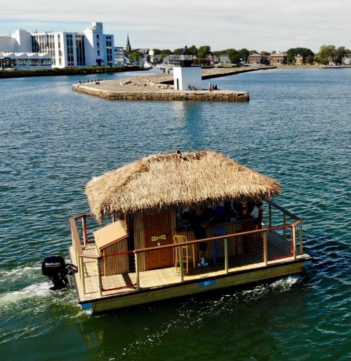 A small floating hut with a thatched roof glides on water, with a shoreline and buildings in the background.