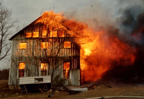 A barn engulfed in flames, with fire visible through the windows and smoke billowing into the sky.