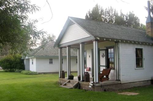 A charming white house with a green roof, featuring a porch and surrounded by a grassy lawn and trees.