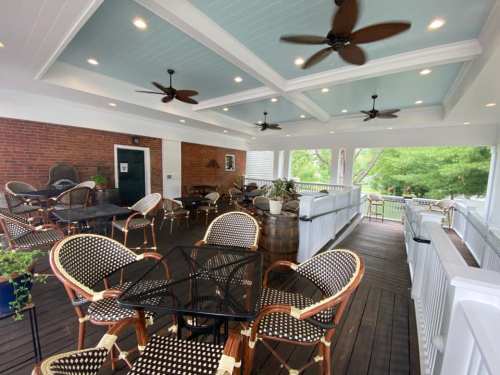 A spacious outdoor patio with ceiling fans, wooden flooring, and tables surrounded by chairs, overlooking greenery.