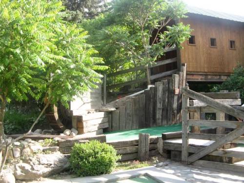 A wooden structure with a water wheel, surrounded by greenery and a pathway leading to a building in the background.