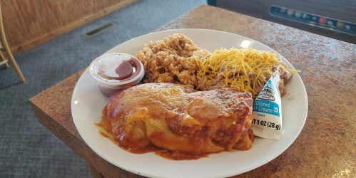 A plate of enchiladas topped with cheese, served with rice, beans, and a side of sauce and sour cream.