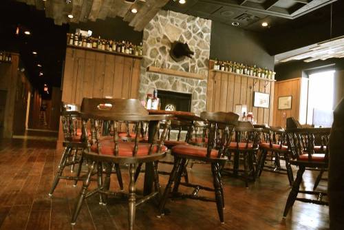 Cozy restaurant interior with wooden tables, chairs, and a stone fireplace, decorated with bottles on shelves.