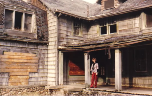 A person stands in front of a weathered, abandoned wooden house with peeling paint and a dilapidated porch.