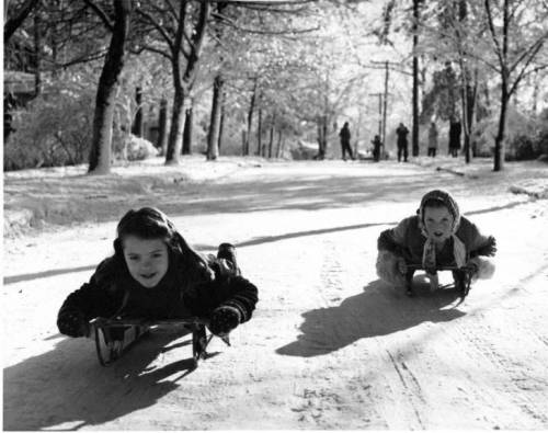 Two girls sledding down a snowy path, surrounded by trees and people in the background, in a vintage black-and-white photo.
