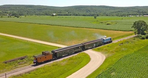 A vintage train travels along a winding track through lush green fields under a clear blue sky.