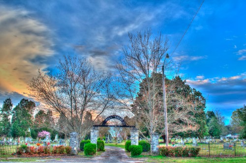 A cemetery entrance with a stone archway, surrounded by trees and colorful flowers under a cloudy sky.