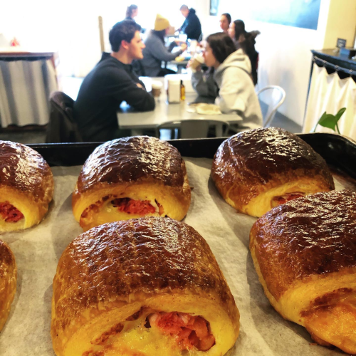 Freshly baked pastries in the foreground with people enjoying meals at tables in a cozy café setting.