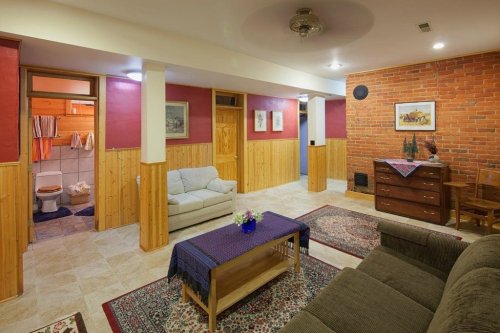 Cozy basement living area with a sofa, armchair, wooden accents, and a brick wall, featuring a small dining table and bathroom.