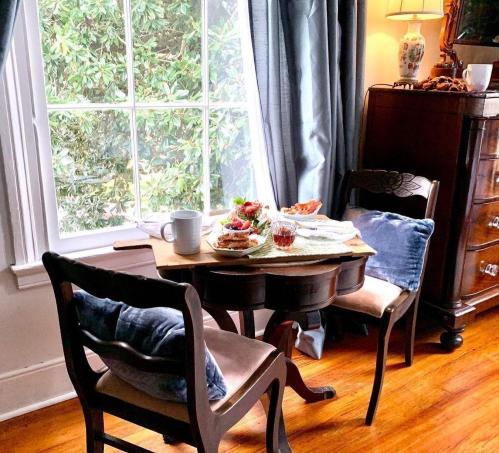 A cozy dining setup with two chairs, a small table, and a spread of food by a window with soft curtains.