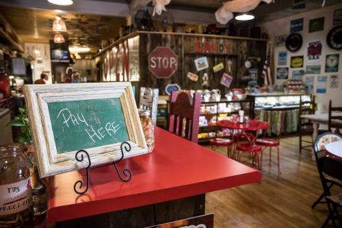 A cozy café interior with a red table, chalkboard sign saying "Pay Here," and vintage decor on the walls.