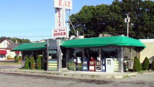A classic diner with a green awning, neon sign, and newspaper stands outside, set in a suburban area.