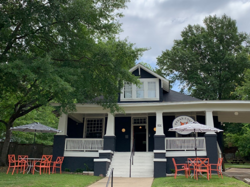 A dark blue house with white trim, featuring a porch and outdoor seating with red chairs and striped umbrellas.